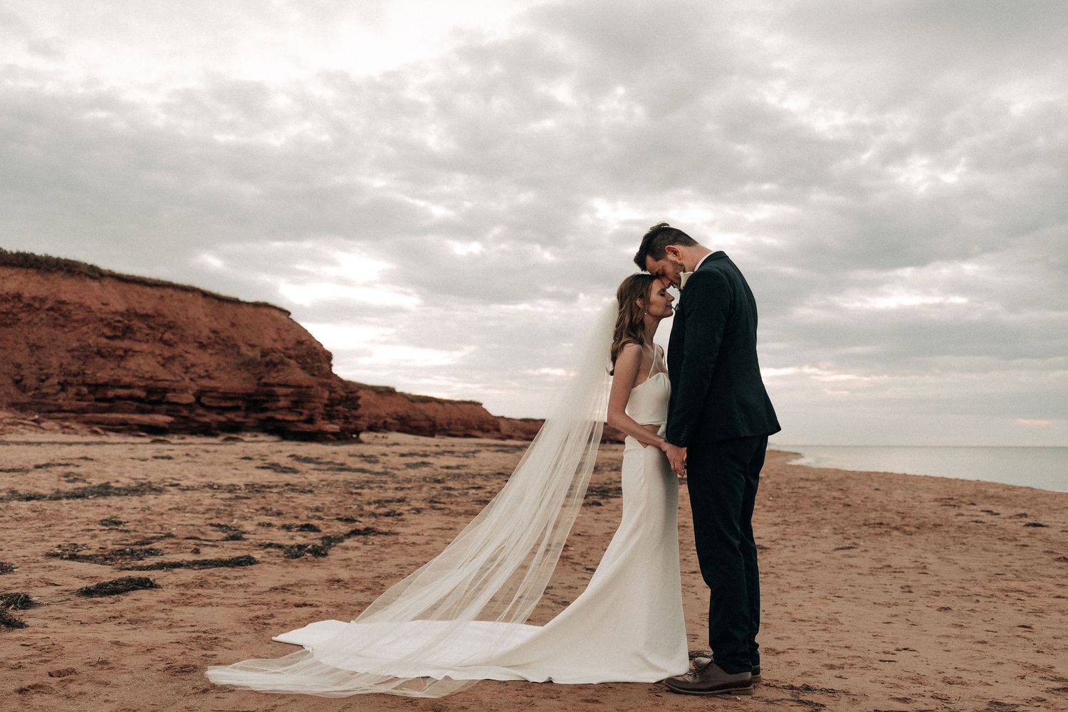 Wedding couple embracing on beach with dramatic red cliffs and cloudy sky.