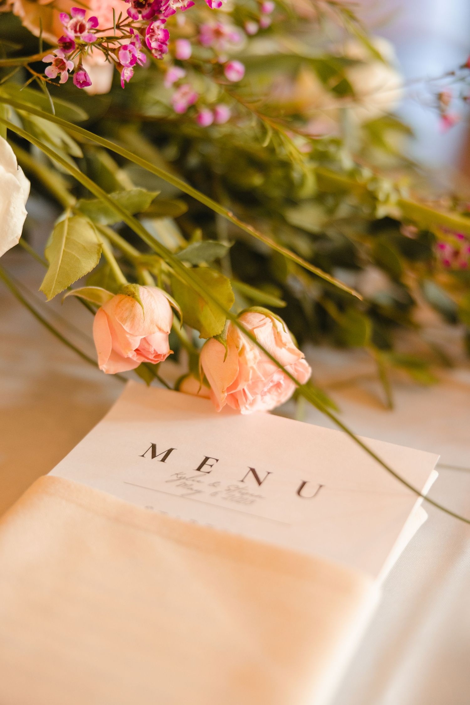 Close-up of a elegant wedding menu card surrounded by soft pink roses and colorful floral arrangements.