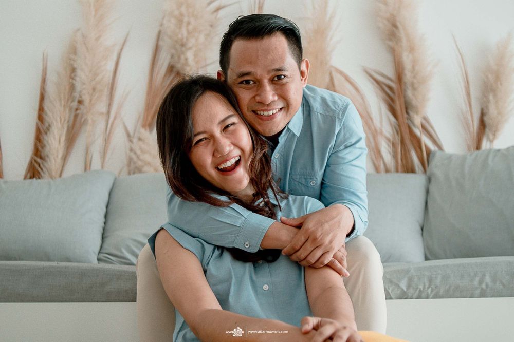 A couple in matching blue outfits shares a fun embrace on their gray sofa with pampas grass decor.