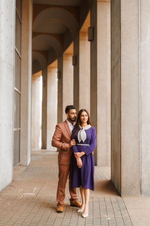 A couple poses elegantly between cream-colored columns in coordinated formal attire.