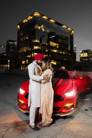 Couple embracing by red sports car at night with illuminated office building in background.