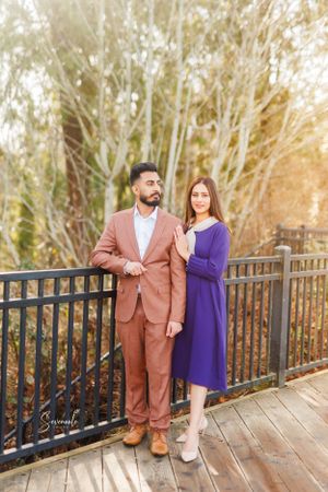 A couple in coordinating purple dress and brown suit pose on a wooden deck with trees in background.