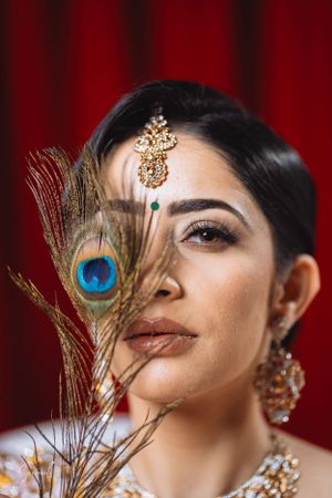 Artistic bridal portrait with peacock feather and traditional gold jewelry against red backdrop.