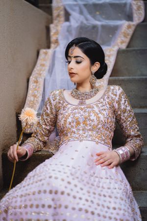 South Asian bride in embellished pink lehenga reclines on concrete stairs with dried flower accent.