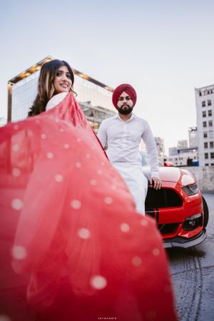 Couple posing with red sports car against urban backdrop with red flowing fabric.