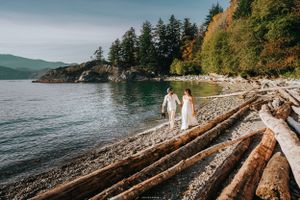 A couple in white attire walks along driftwood logs on a scenic coastal beach.