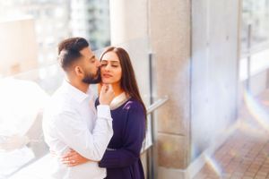 A romantic couple embraces intimately against a white wall outdoors.