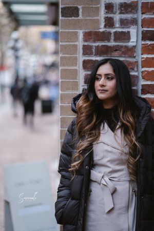 A woman in a black winter coat and beige dress poses against a brick wall on a city street.