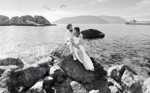 A romantic black and white photo of a couple on coastal rocks with birds flying overhead.