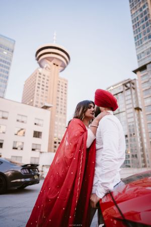 Romantic couple standing by red sports car with Vancouver Lookout tower visible in background.