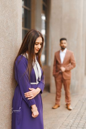 A couple poses in formal wear, with a woman in a purple dress and man in a brown suit against a beige wall.