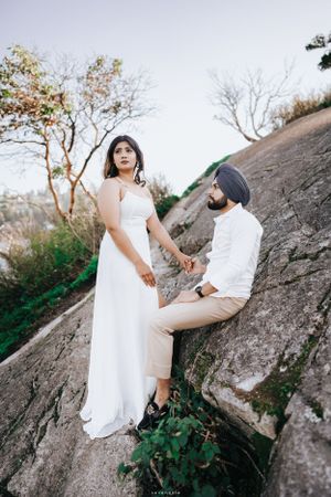 A couple poses dramatically on a rocky cliff edge wearing elegant white attire.
