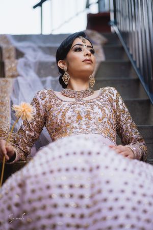 Indian bride in ornate gold sequined dress and luxurious jewelry poses elegantly on stairs with dramatic lighting.