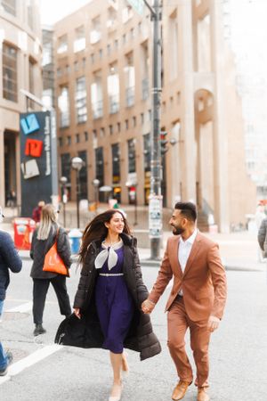 A stylish couple walks hand in hand through a city street, wearing purple dress and brown suit.