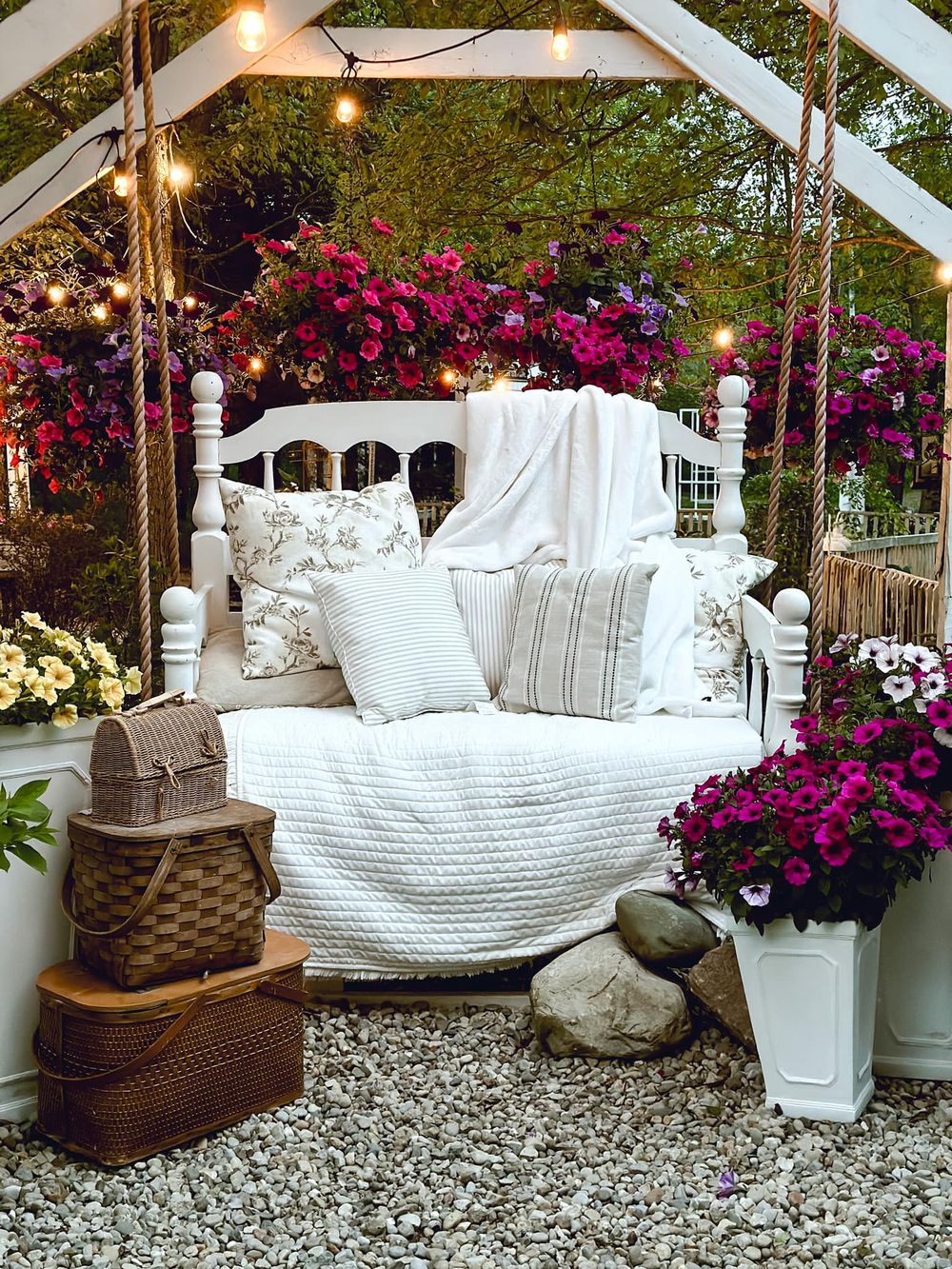 Cozy outdoor bed setting with white linens and vibrant purple flowers under a pergola with string lights.
