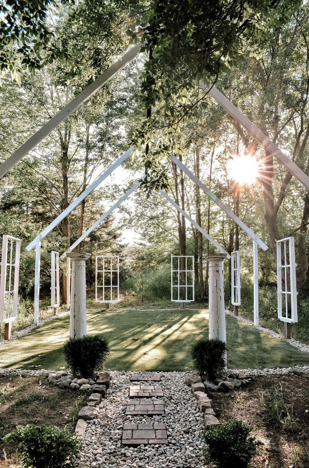 White architectural archway with hanging windows catching sunbeams through a forested garden path.