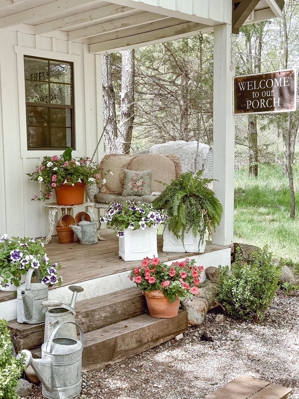 Charming farmhouse porch decorated with potted flowers, ferns, and vintage watering cans.