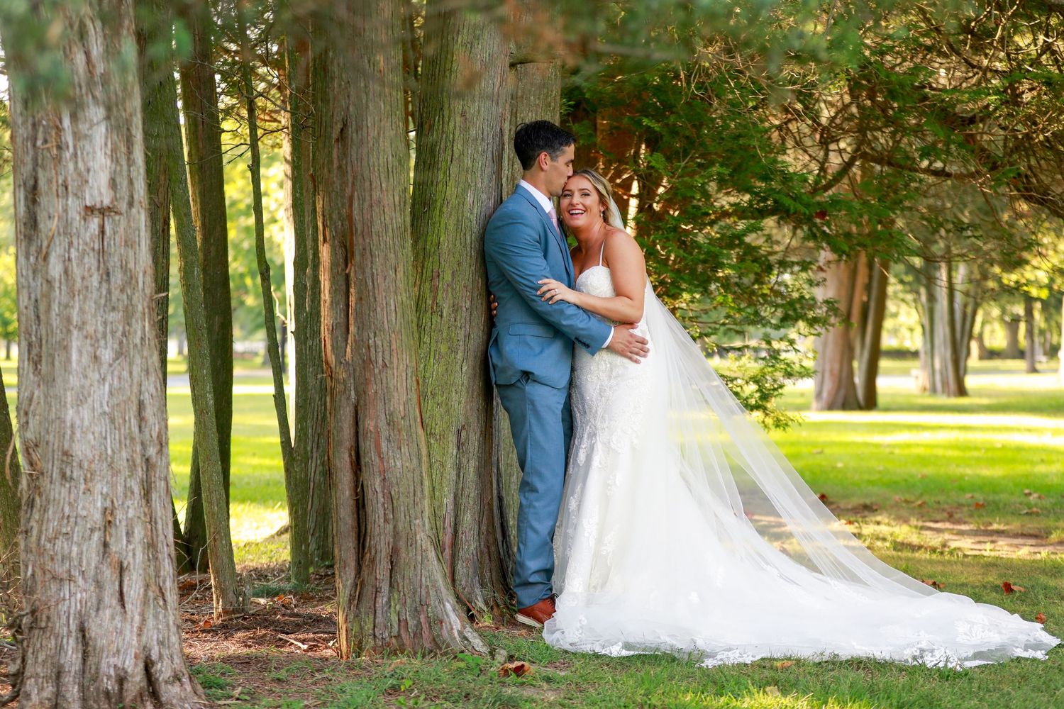 Groom tenderly kisses bride near tall trees in a lush green park setting.