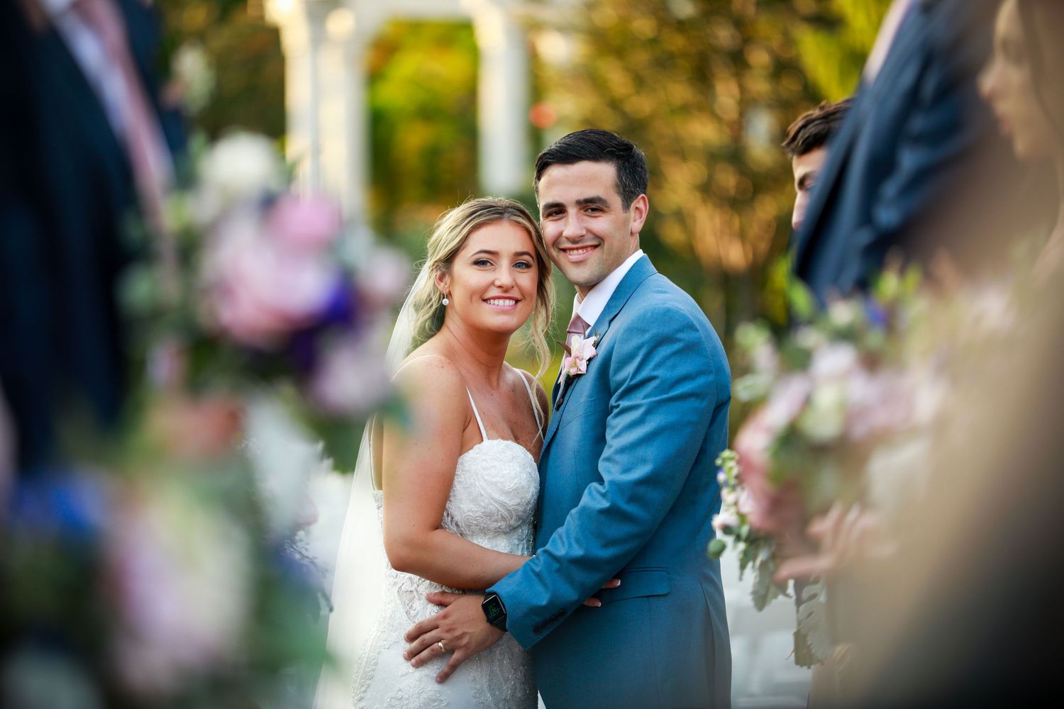 Smiling bride and groom pose together at outdoor ceremony surrounded by pink and purple florals.