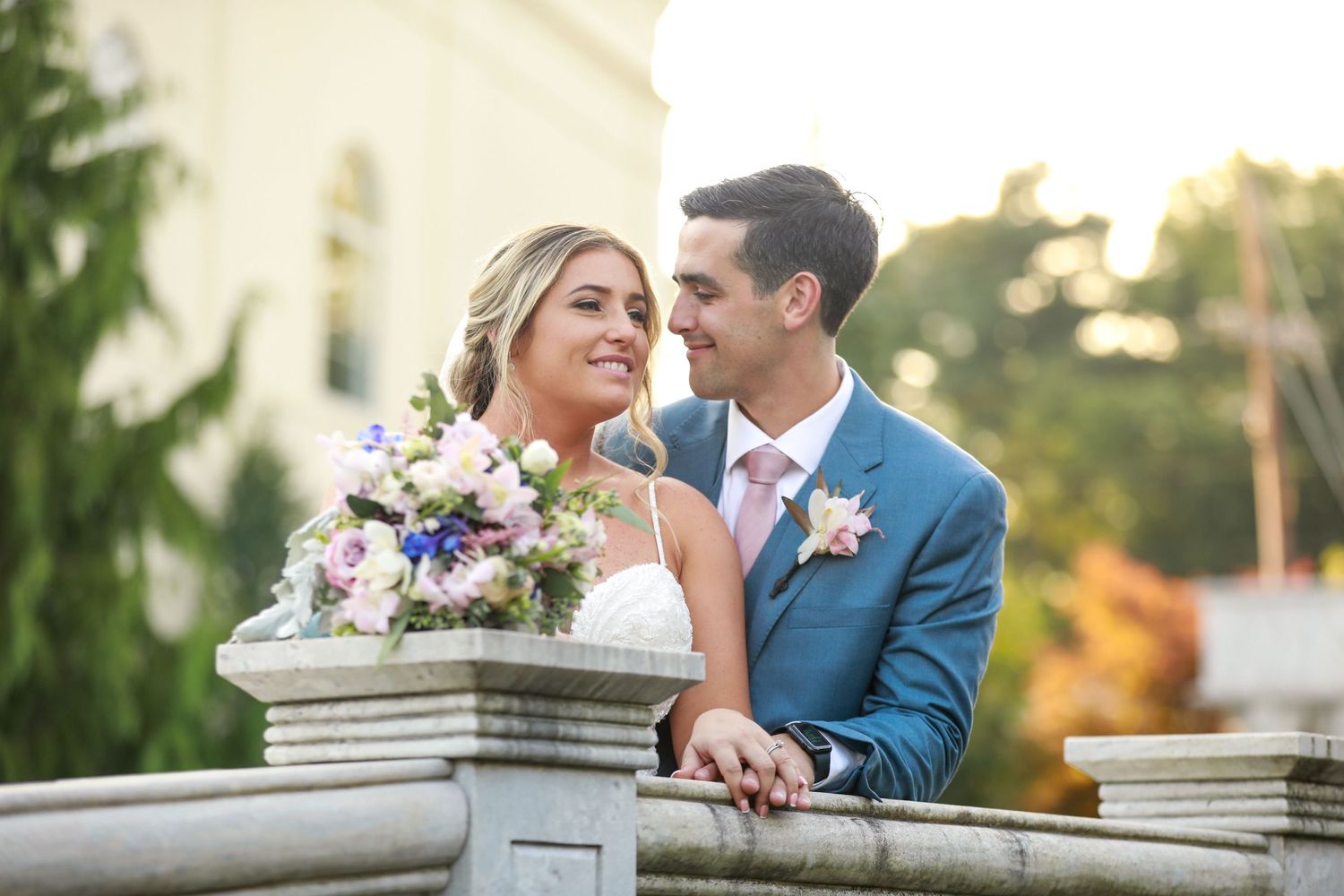 Bride and groom share a joyful moment on a stone balcony with a colorful bouquet.