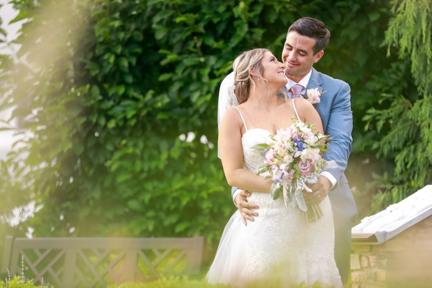 Bride holding bouquet and groom embracing outdoors in a lush green garden on their wedding day.
