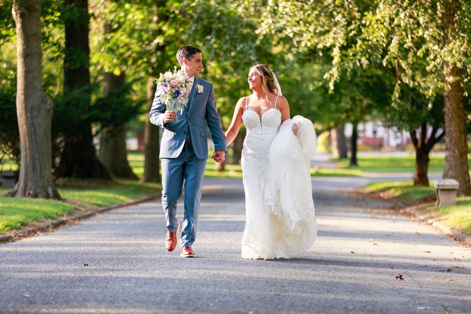 Bride and groom walking hand in hand down a tree-lined path on their wedding day.