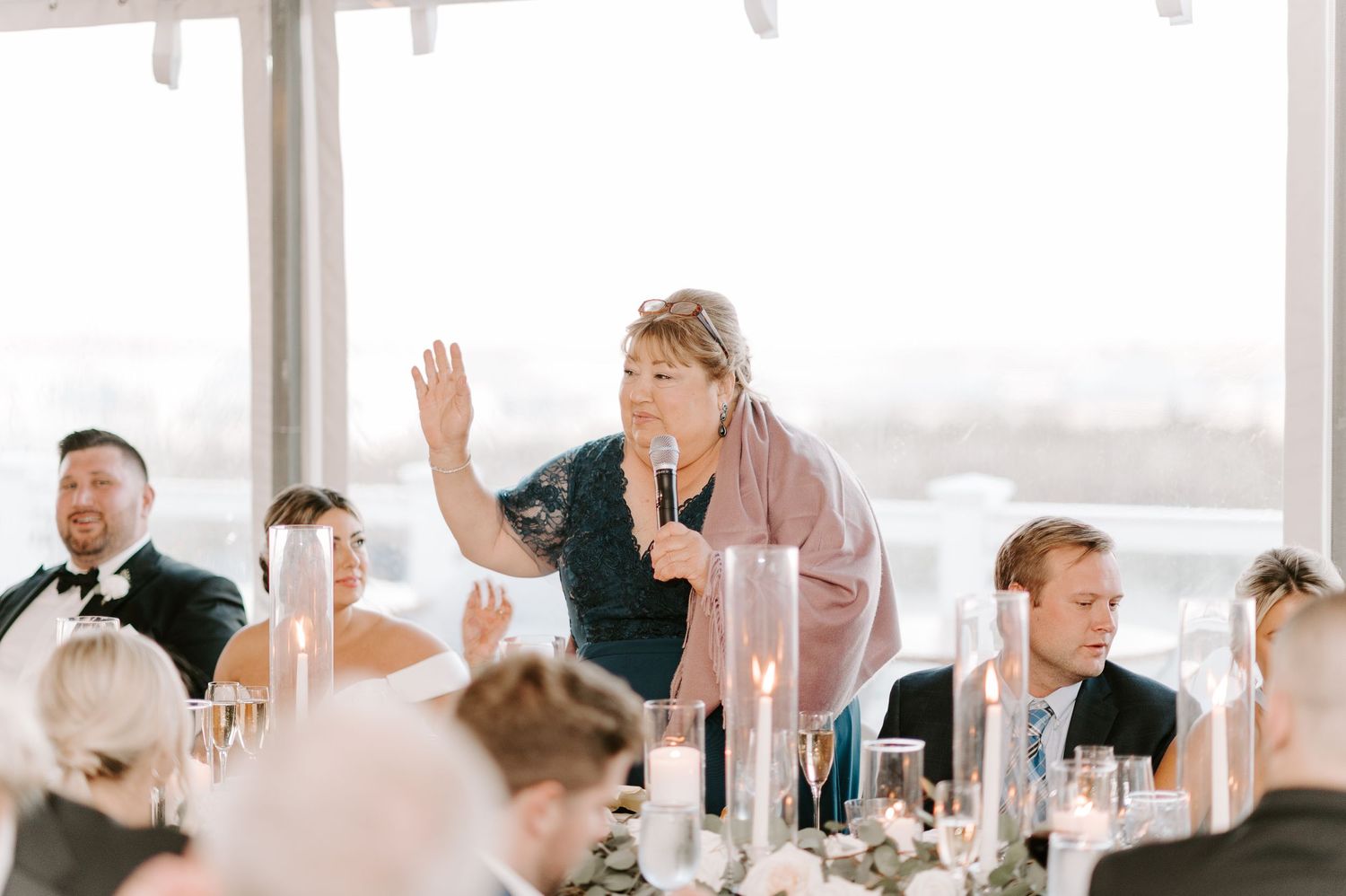 Wedding guests react with joy during speeches at a sunlit reception venue.