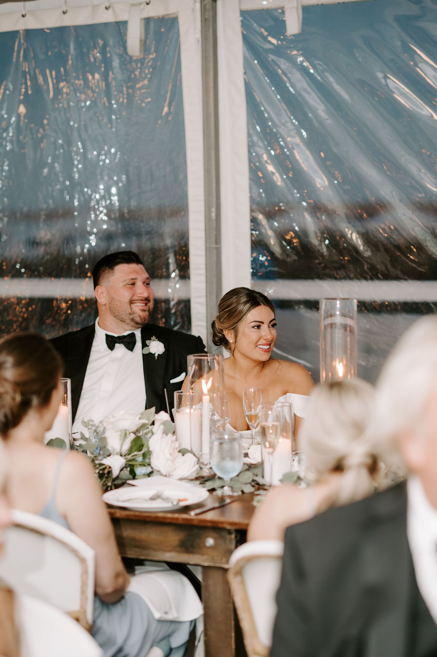 Wedding party members share laughs during reception dinner in a tented waterfront venue.