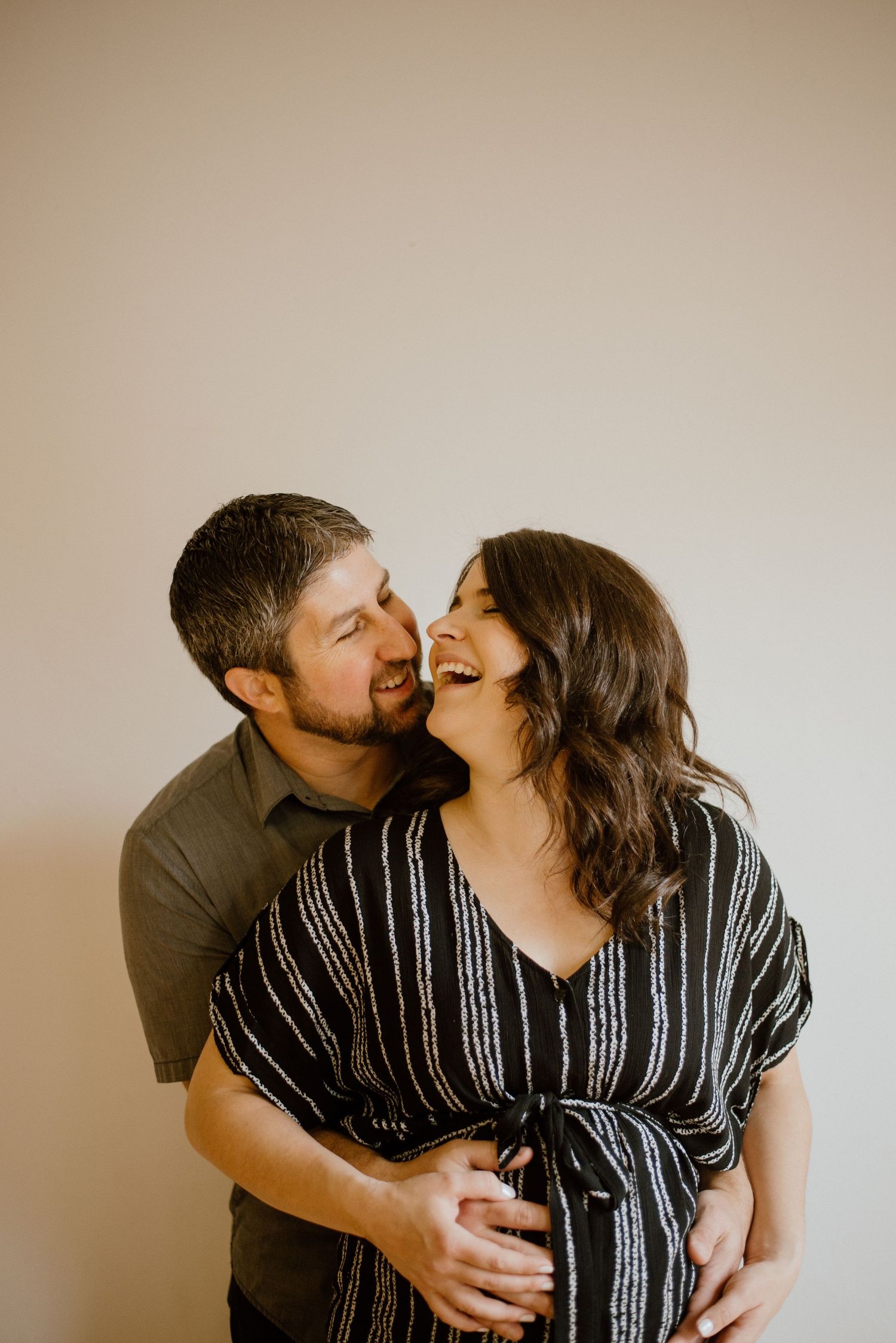 Two people share an intimate laughing moment against a beige background.