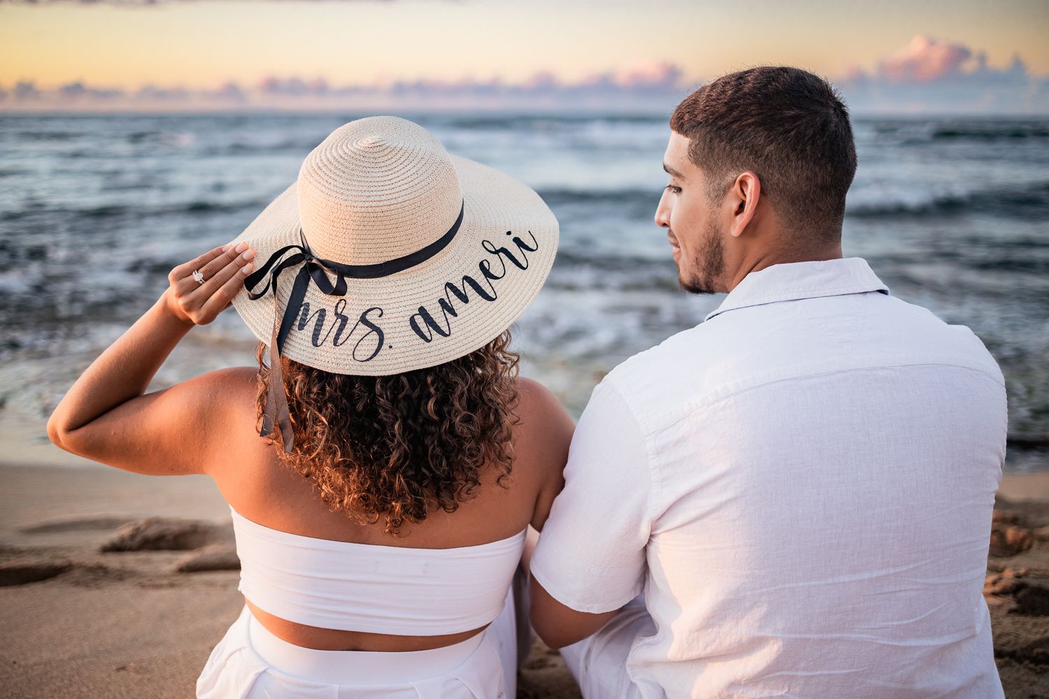 Couple sits on beach at sunset wearing white, with woman in "Mrs" sun hat looking at horizon.