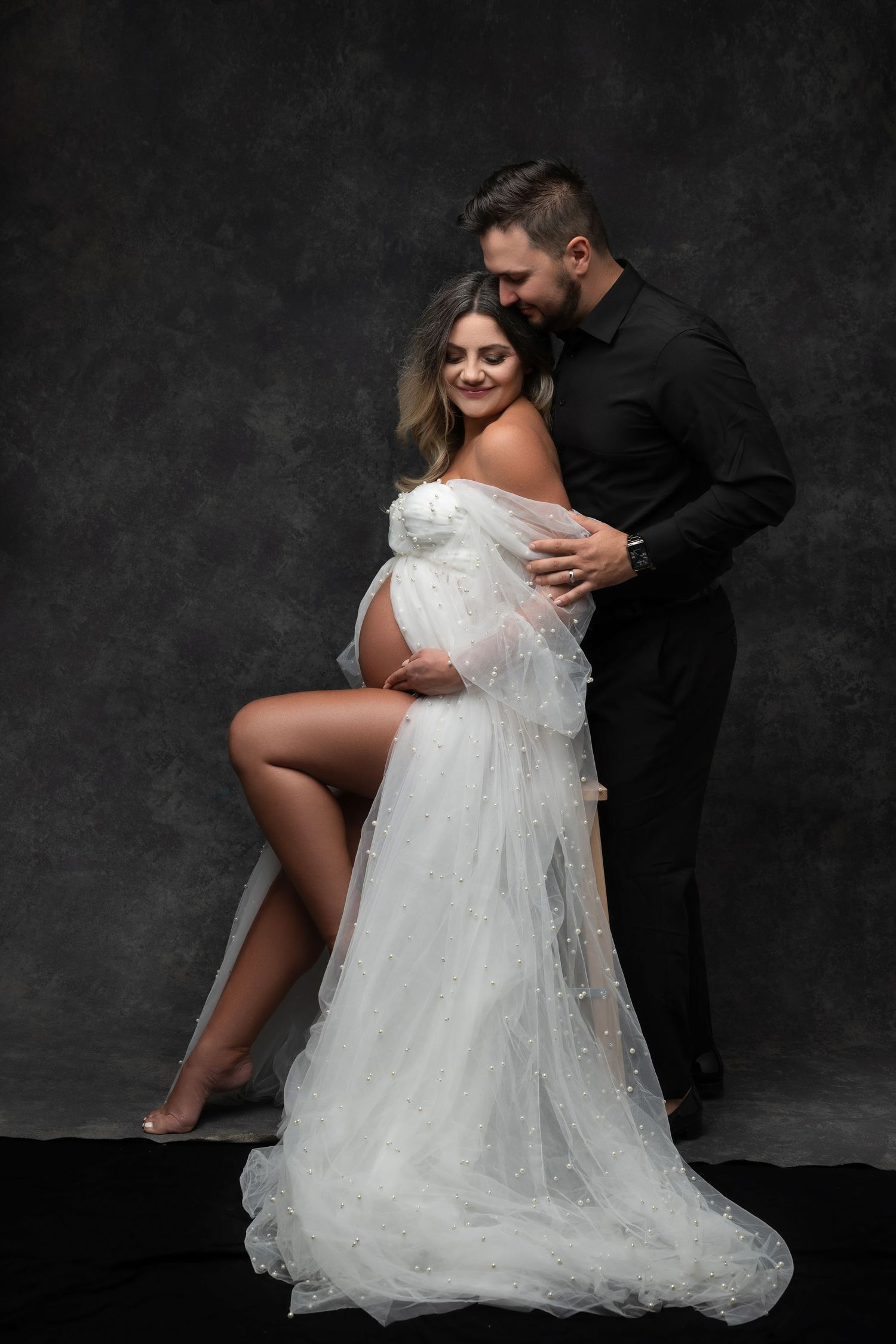 Romantic maternity portrait with couple in white gown and black attire against dark background.