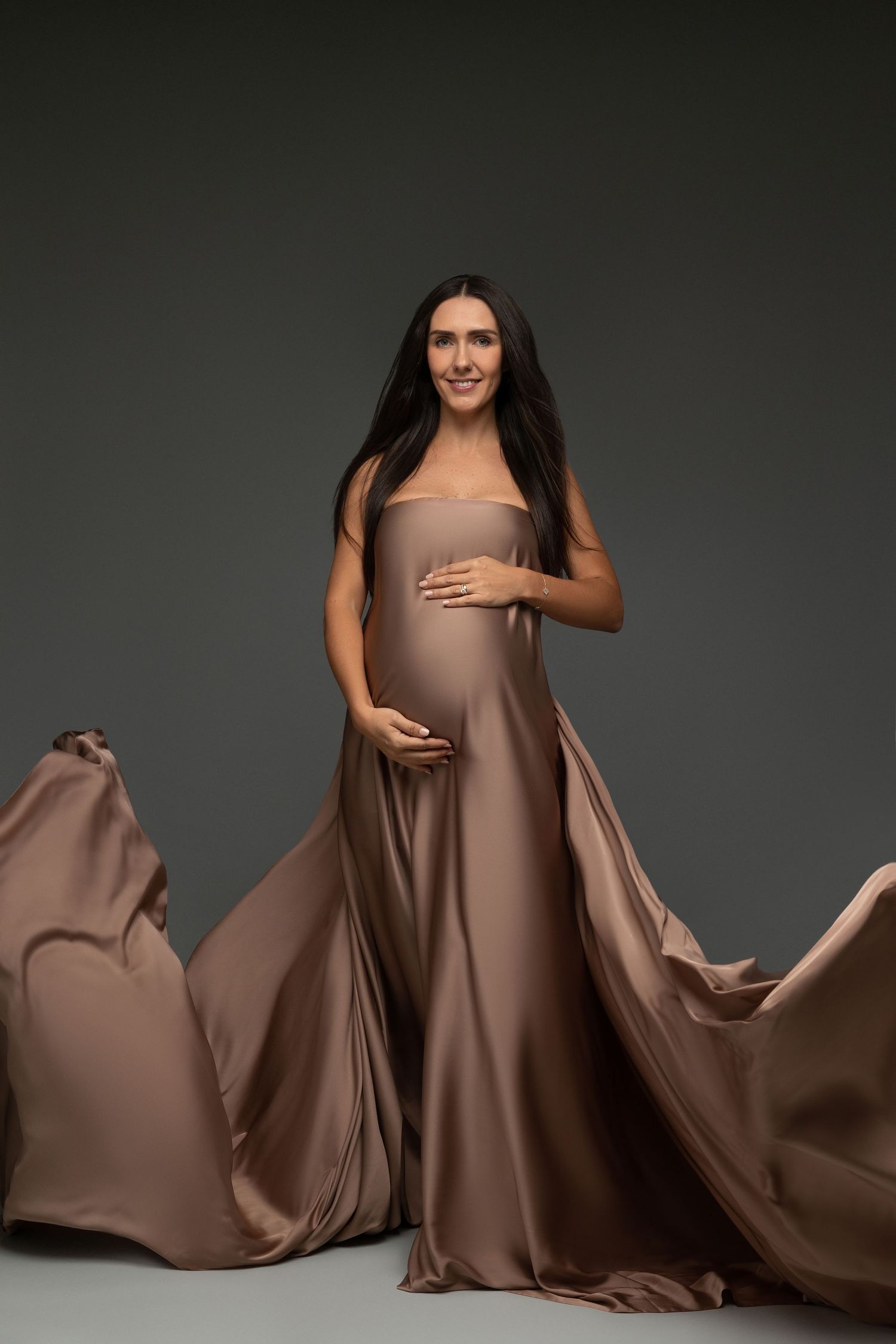 Elegant maternity photo featuring flowing mocha-colored dress against dark background.