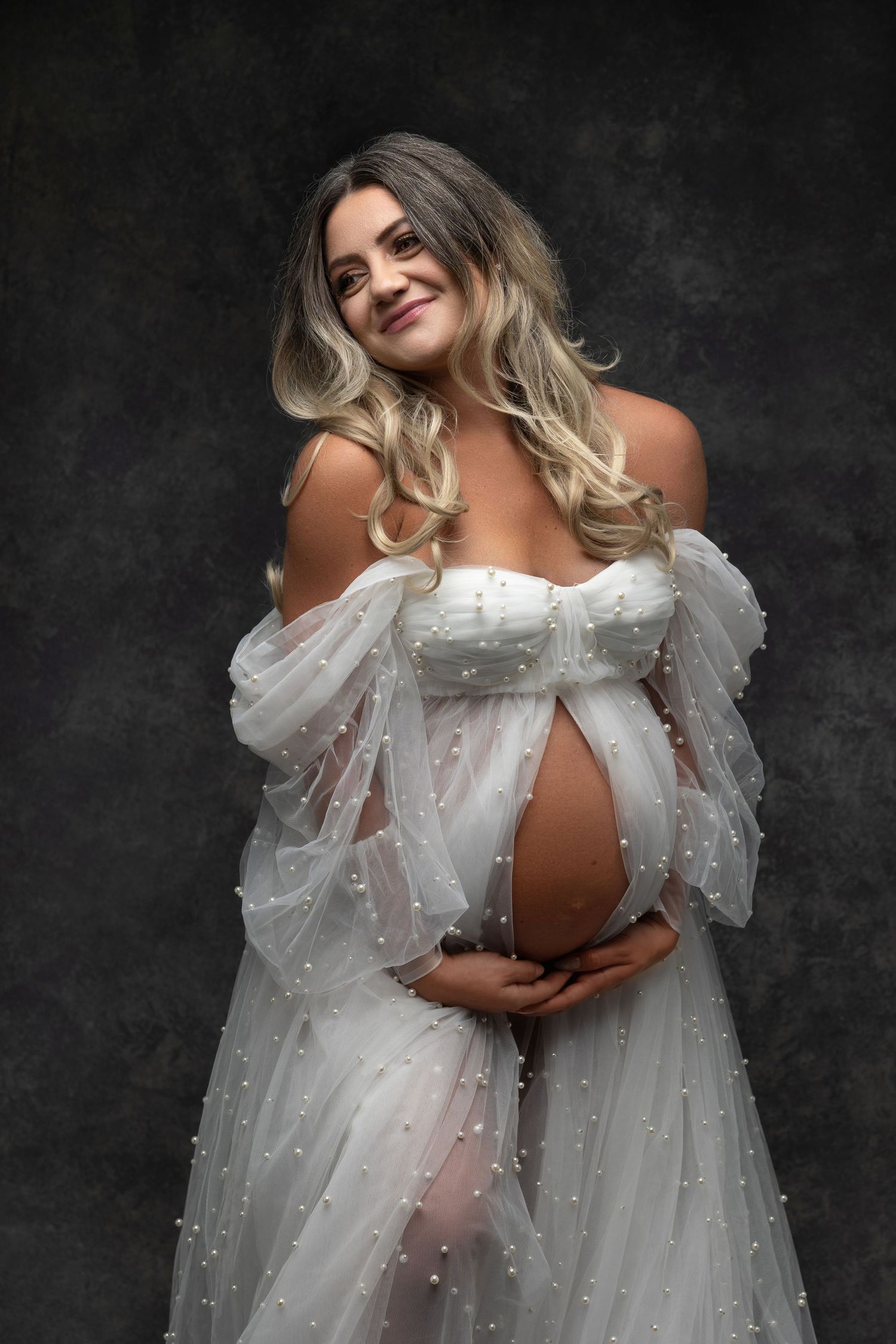 Ethereal maternity portrait in off-shoulder white pearl-adorned gown against dark backdrop.