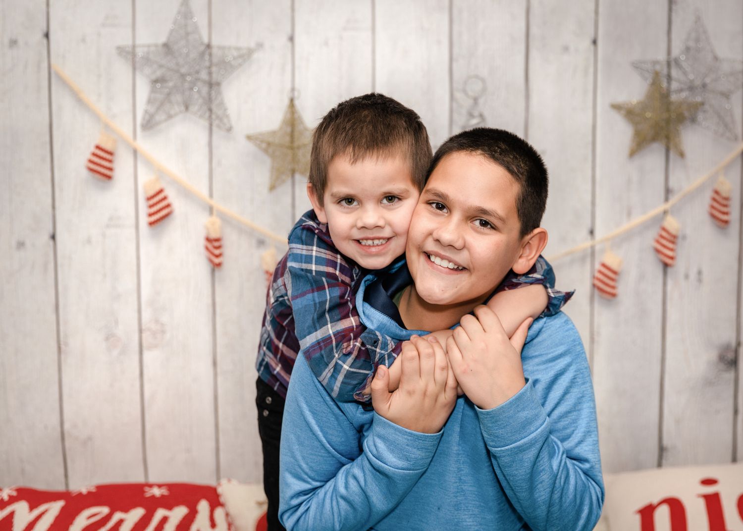 Two boys hugging and smiling in front of a rustic Christmas holiday backdrop with stars.