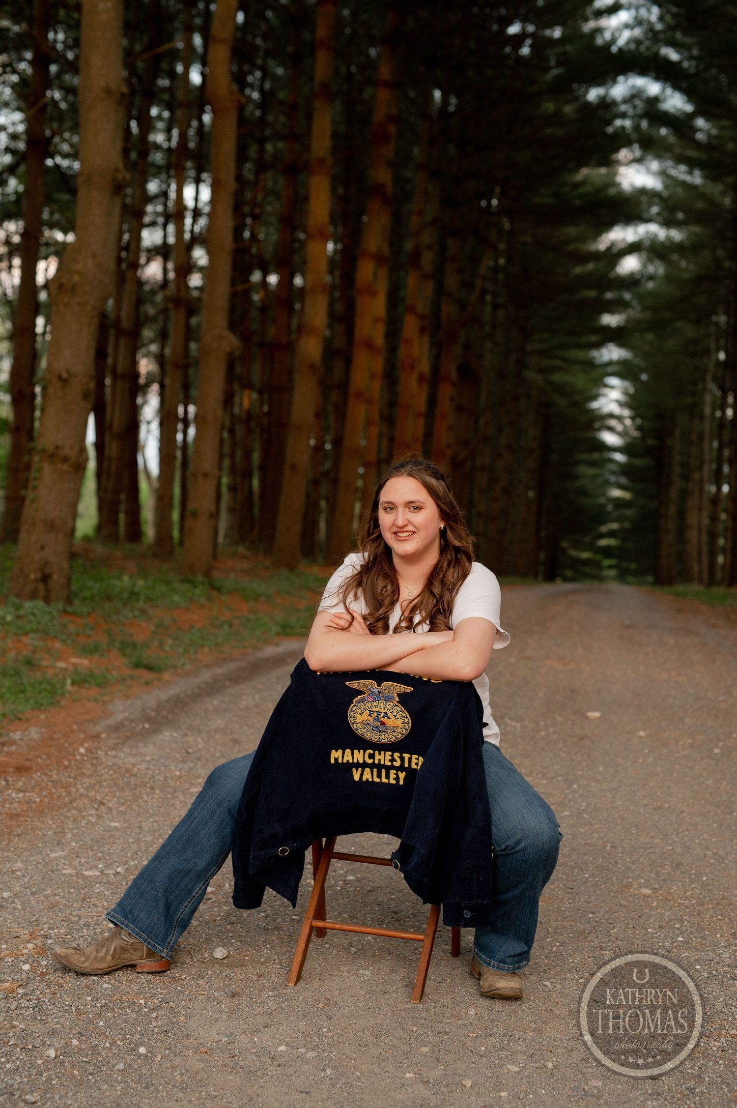 A casual seated portrait on a wooden chair along a tree-lined dirt path.