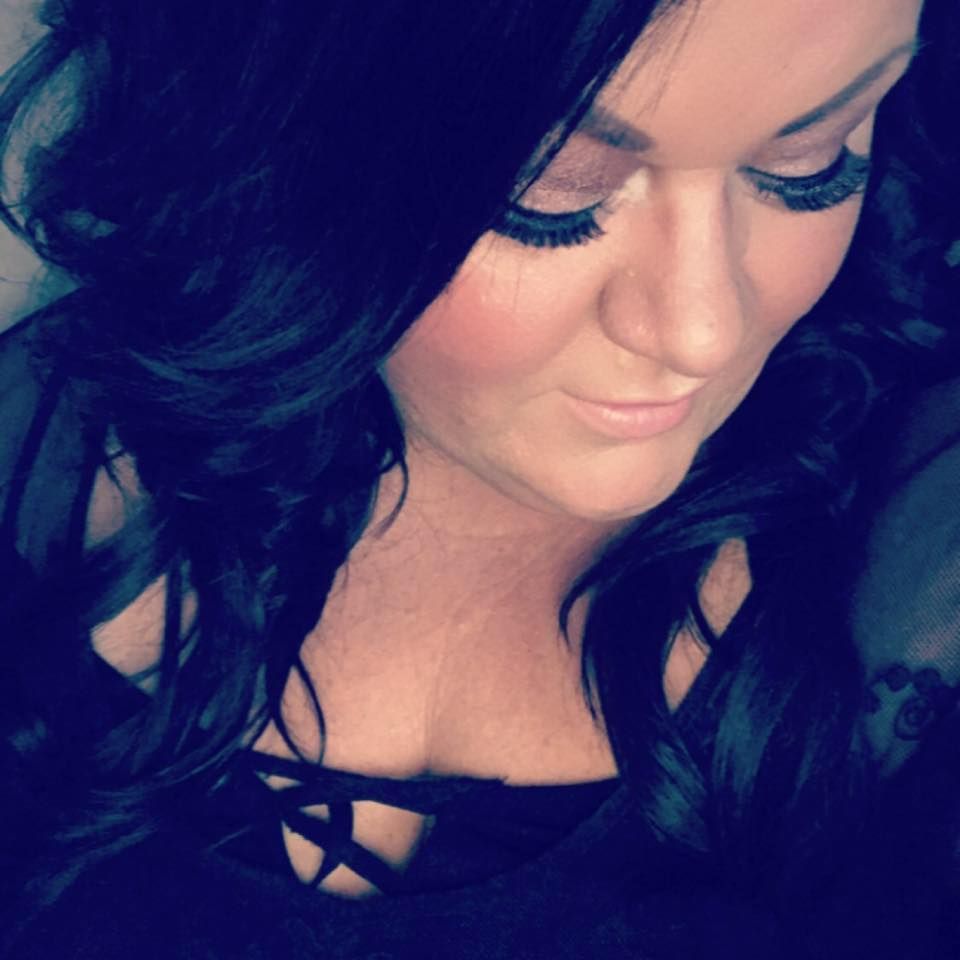 A woman with long dark curly hair and dramatic lashes looks down in a black top.