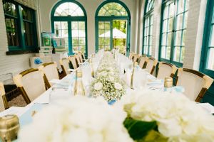 Elegant white baby shower reception table setup in sunlit room with green arched windows.
