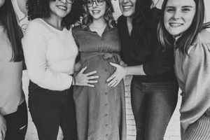 A black and white photo of friends gathered together smiling and placing their hands on a pregnant belly.