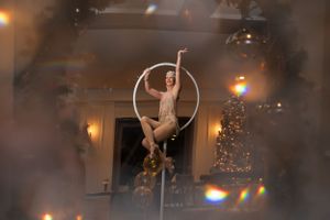 An aerial performer gracefully poses on a hoop against warm lighting and a Christmas tree.