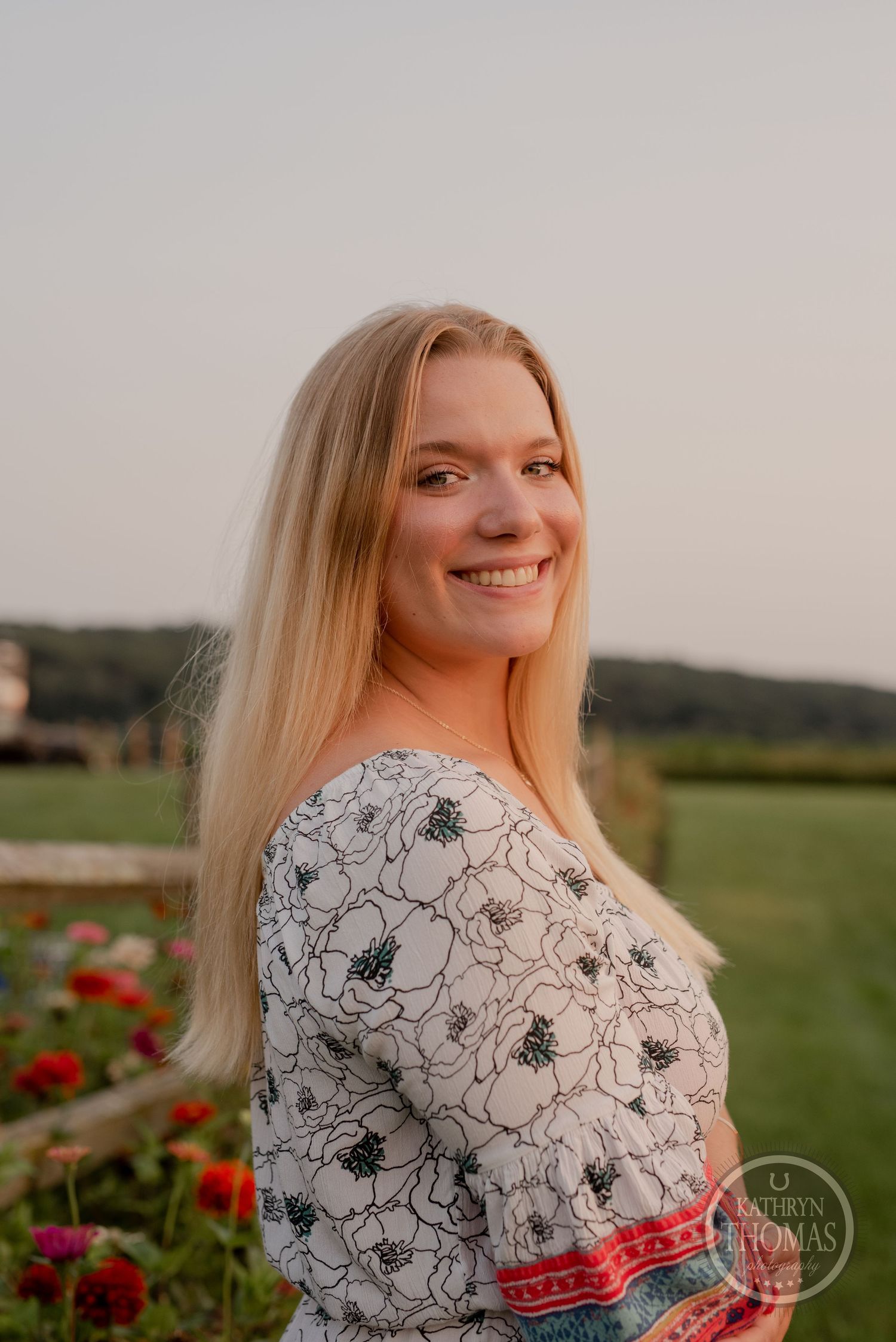 A smiling woman in a floral print romper poses in front of a garden with red flowers at sunset.