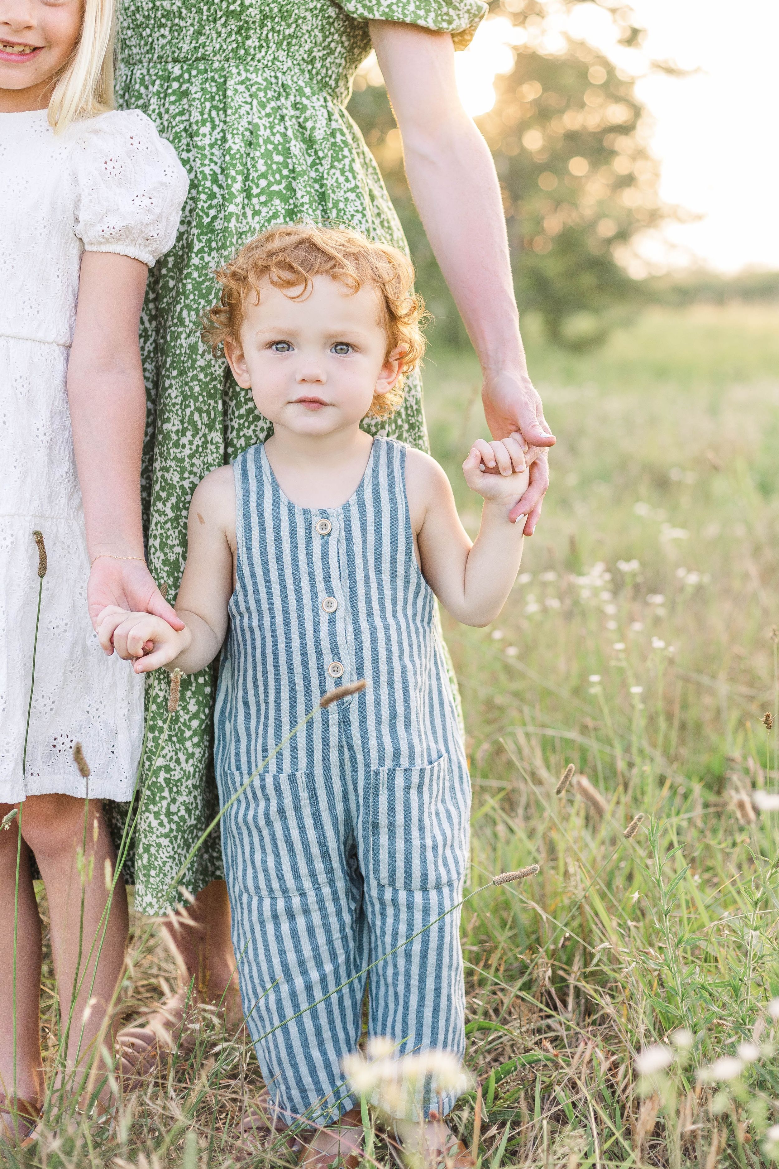 Young child wearing blue overalls standing in a grassy field while holding a parent's hand during an outdoor family photo session.