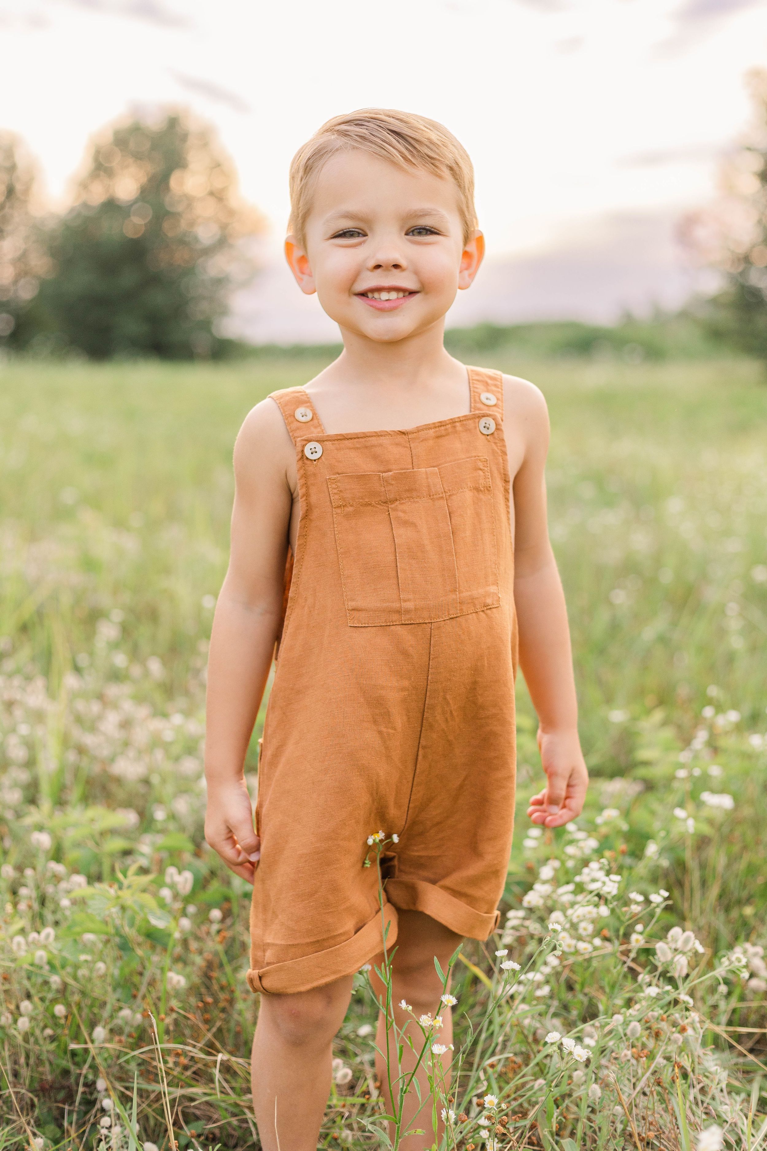 Smiling young child wearing a rust-colored outfit standing in a grassy field during a golden hour outdoor portrait session.