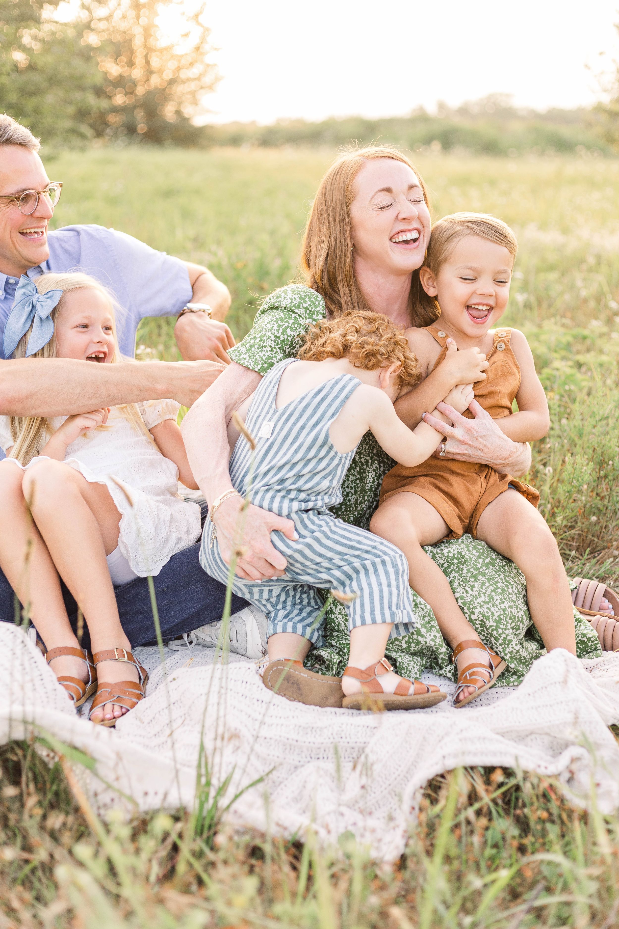 Young child wearing blue overalls standing in a grassy field while holding a parent's hand during an outdoor family photo session.