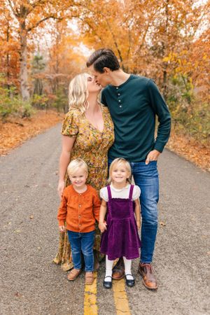 Father standing with his two young children on a pathway surrounded by vibrant orange autumn trees during a fall portrait session.