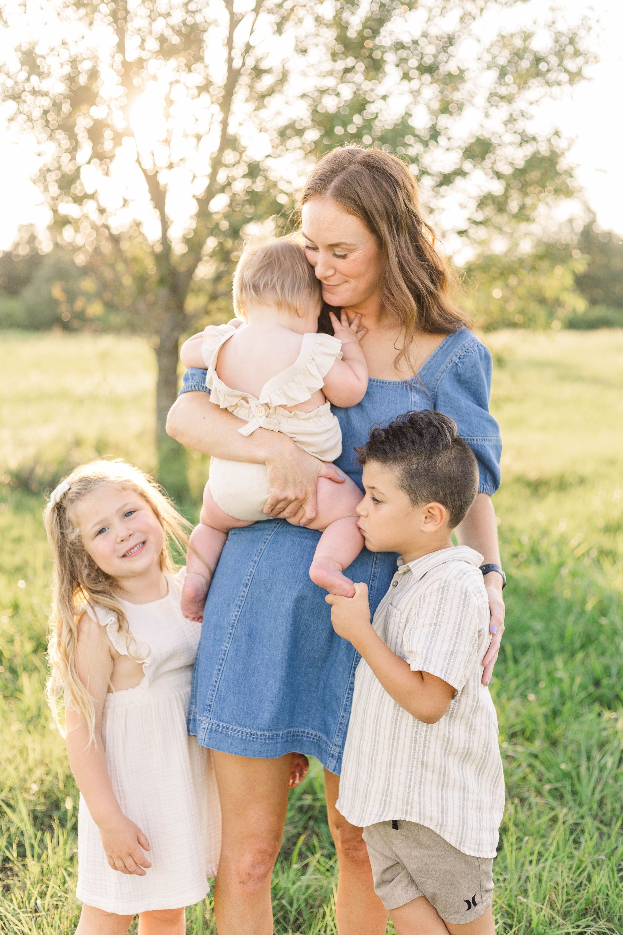 Mother holding her baby while standing with two young children in a sunlit field during an outdoor family portrait session.