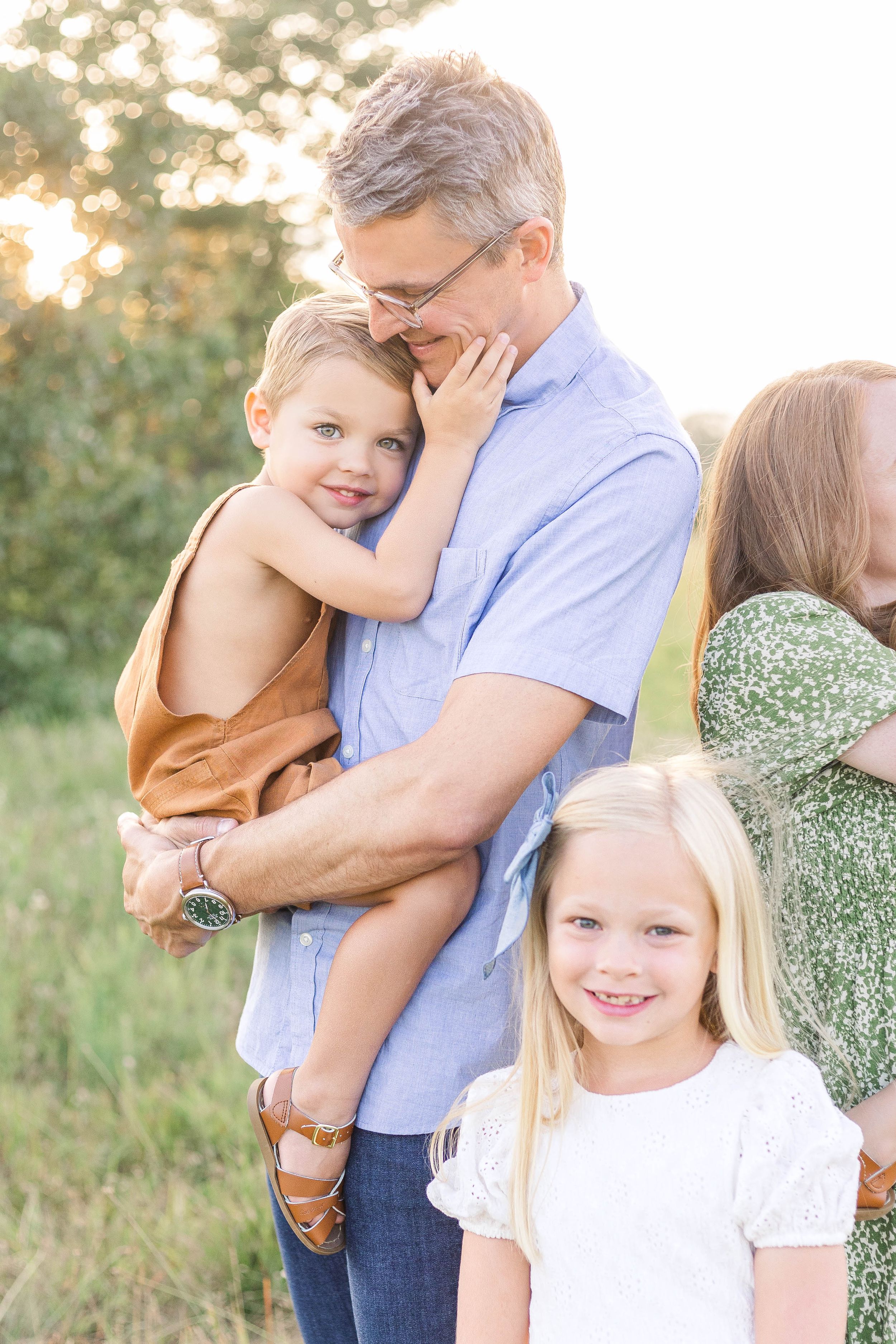 Father holding his young child while standing with his daughters outdoors during a warm, natural light family portrait session.