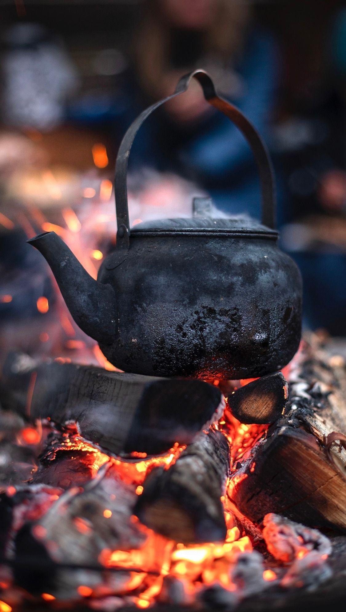 Black cast iron kettle heating over glowing campfire embers with sparks flying in the dark.