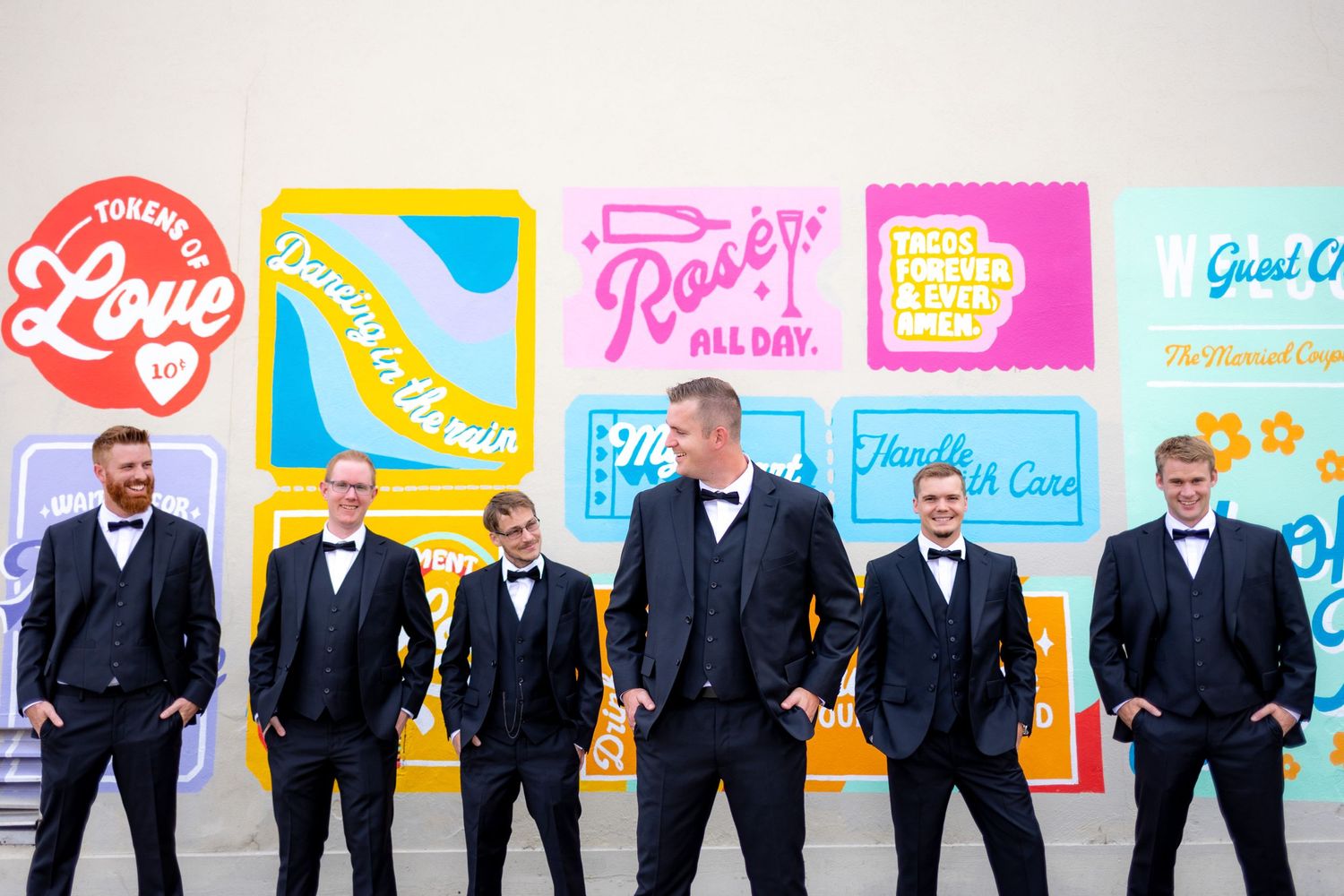 Groomsmen in formal attire pose against colorful wall art mural with retro style designs.