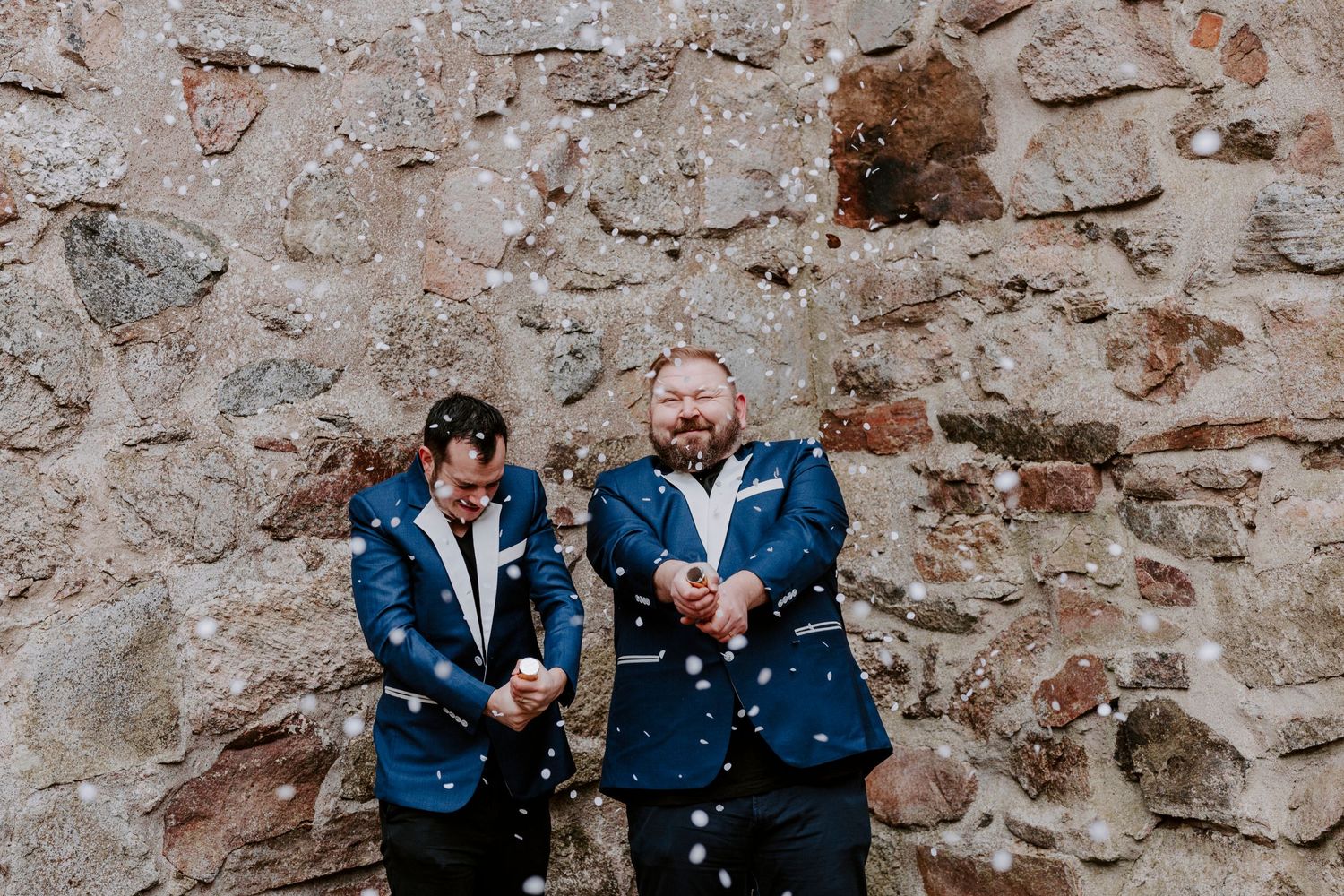 Two people in matching blue suits laugh together against a rustic stone wall.