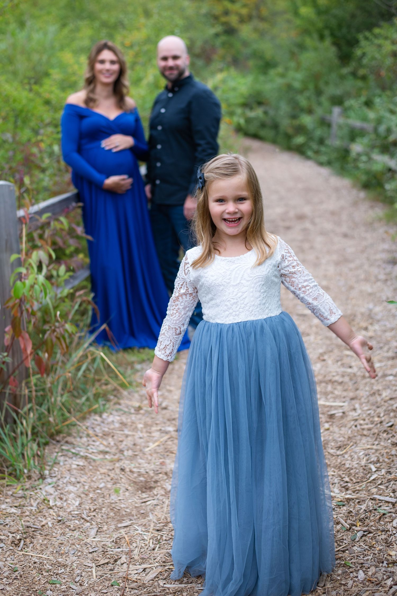 Young girl in blue tulle dress poses joyfully on nature path with parents in background.